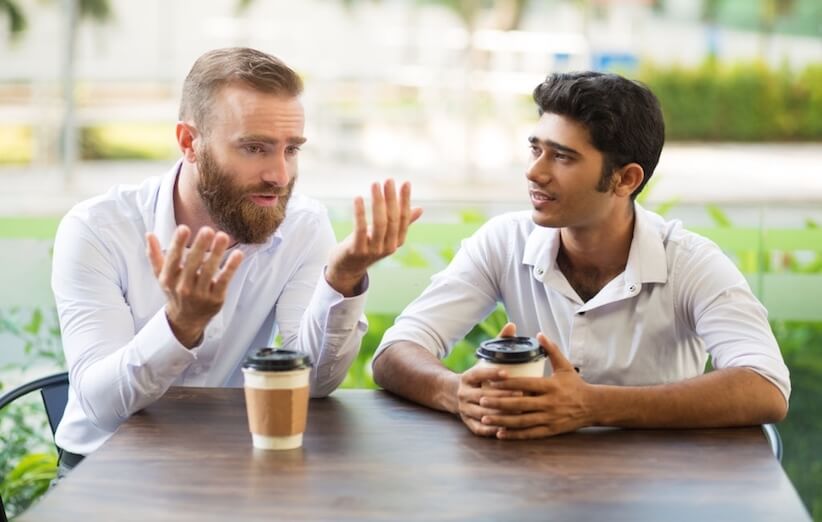 دو مرد در کافه‌ای در حال صحبت کردن با یکدیگر هستند. هر دو لیوان قهوه دارند.