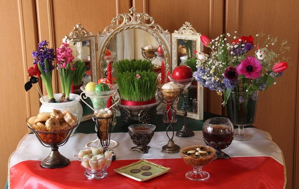 هفت سین عید 1402 Tabletop with Haft-seen elements for Nowruz: sonbol (hyacinth), sabzeh (grass), seeb (apple), somaq (sumac powder), seer (garlic), serkeh (vinegar), goldfishes, flowers hyacinths, coins, burning candles, painted eggs and mirror