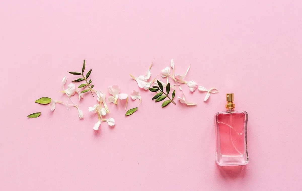 شیشه عطر در پس زمینه صورتی با گلبرگ‌های سفید و برگ‌های سبز 