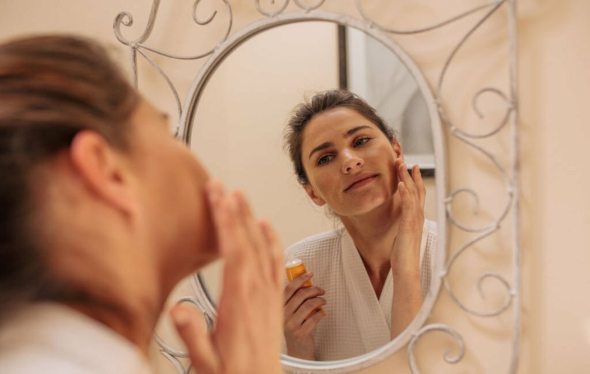 یک زن در جلوی آینه که بهترین ضد افتاب بی رنگ برای پوست جوش دار را روی صورت خود می‌زند 