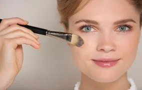 یک زن جوان که کرم پودر را با برس آرایشی روی صورت خود می‌زند