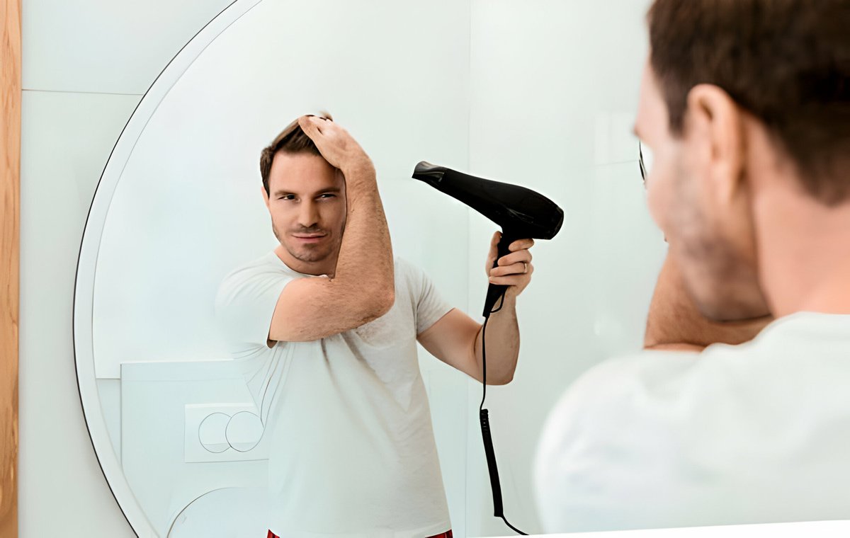 یک مرد جوان که روبه‌روی آینه ایستاده است و موهایش را سشوار می‌کشد