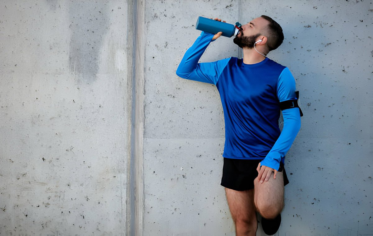 یک مرد جوان به دیوار تکیه داده است و نوشیدنی ورزشی می‌خورد