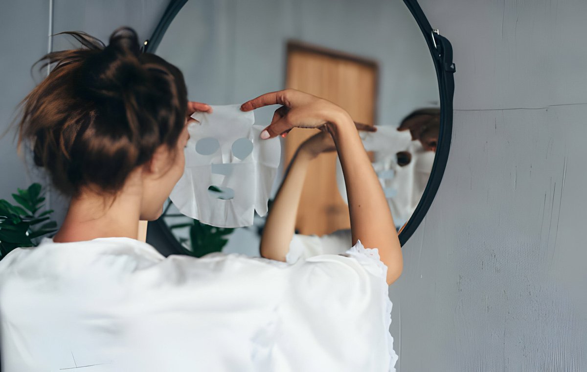 یک زن که جلوی آینه ایستاده است و روی صورت خود ماسک ورقه‌ای می‌گذارد