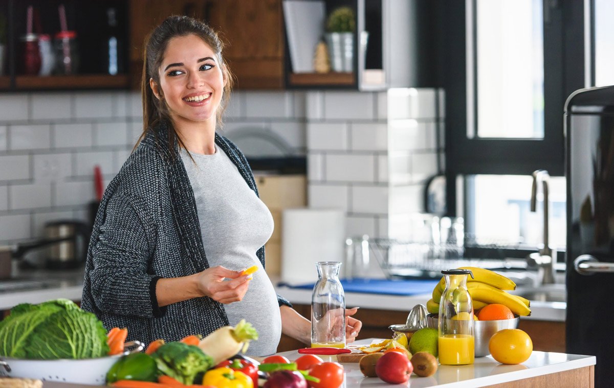 یک زن باردار در آشپزخانه که مقدار زیادی میوه و سبزی روبه‌روی او دیده می‌شود.