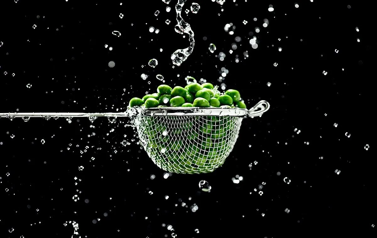 یک سبد فلزی پر از نخود فرنگی که در حال شسته شدن در یک بک گراند مشکی است