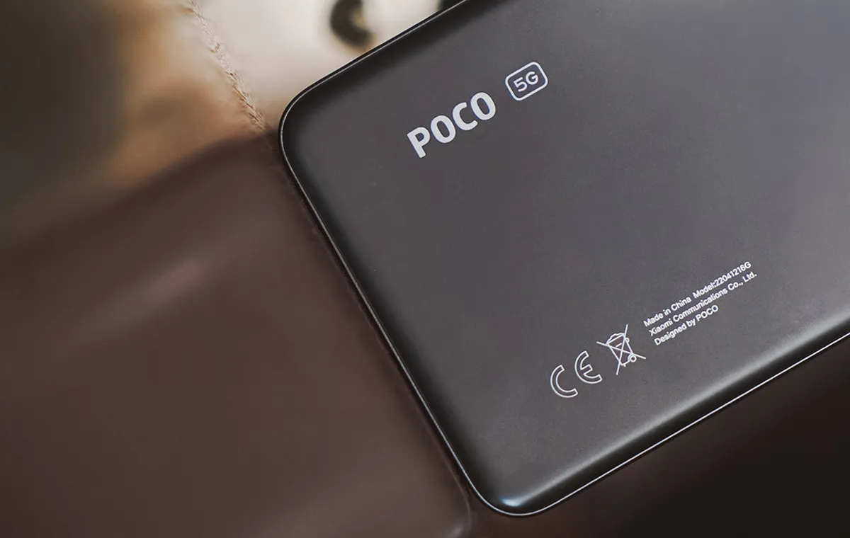 نیمه‌ی پایینی یک گوشی مشکی پوکو روی میزی نیمه‌شفاف قرار دارد که تصویر گوشی را منعکس کرده است